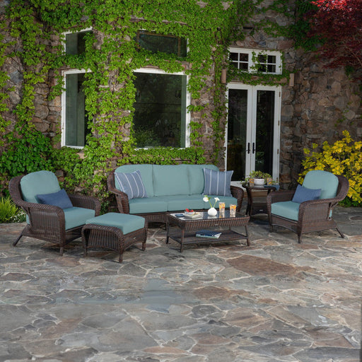 The Tortuga Outdoor Sea Pines 6-Piece Wicker Sofa Set in Java, featuring blue cushions, a sofa, two armchairs, an ottoman, and a coffee table, is arranged on a stone patio for stylish outdoor living.