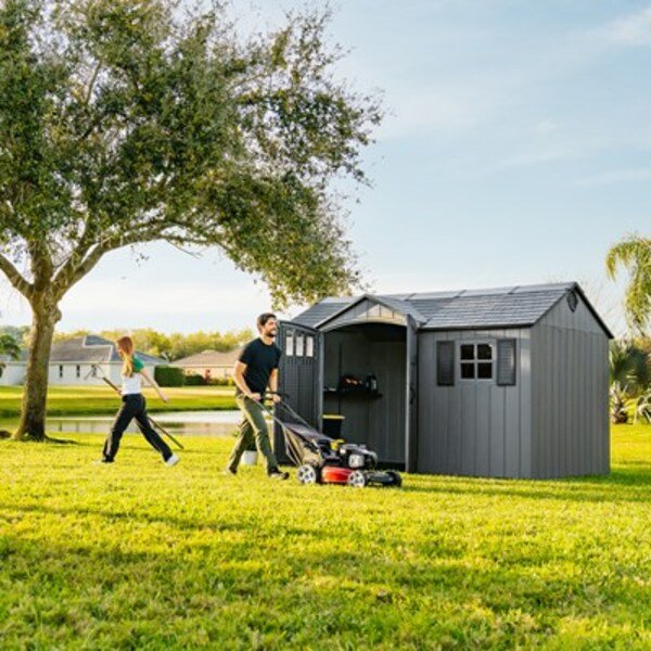 Man with a lawn mower and woman near the Lifetime 12.5x8 ft. storage shed with open door, lake in the background.