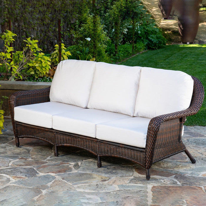 The Tortuga Outdoor Sea Pines 6-Piece Outdoor Wicker Sofa Set in Java, featuring white cushions, sits on a stone patio amid greenery and lawn—perfect for relaxing with stylish Tortuga Outdoor furniture.