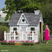 Little Cottage Co. 4x6 Victorian Playhouse with gray shingles, white trim, and flower boxes, featuring a girl in a pink dress smiling outside the front porch.