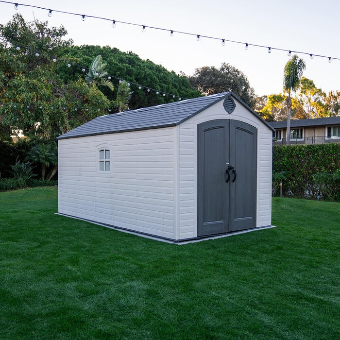 Side view of the Lifetime 8 Ft. x 15 Ft. Outdoor Storage Shed on a green lawn with garden and string lights in the background.