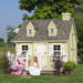 A young girl in a white dress plays outside the pastel green Little Cottage Co. Cape Cod Playhouse featuring a white porch, dormer windows, and flower boxes.