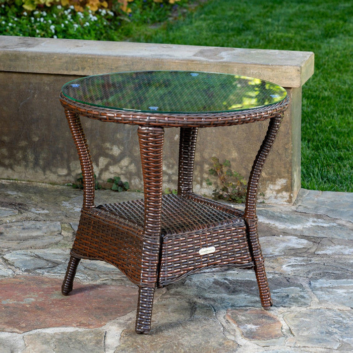 The Tortuga Outdoor Sea Pines 6-Piece Outdoor Wicker Sofa Set - Java by Tortuga Outdoor includes a round wicker side table with a glass top and lower shelf, shown here on a stone patio with grass and a stone wall in the background.