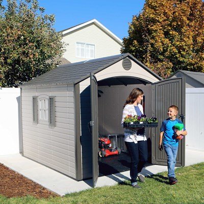 Mother and son carrying plants near the Lifetime 8x10 ft. storage shed with open doors and mower visible inside.
