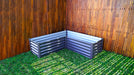 Absco The Organic Garden Co 4' x 4' x 1' Metal L Garden Bed assembled and empty, set against a wooden fence and grass background.