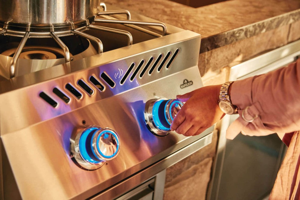 A close-up of a hand adjusting the red control knob of the Napoleon Grills Burner, with a blue flame visible under the pot.