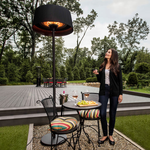 A woman stands on an elegant wooden deck next to a table with snacks and wine glasses, enjoying the warmth from the Black Sol Electric Standing Heater.