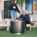 Bonfire Fire Pit in Gunmetal color plus Stand 2.0 placed outside a house with a couple.