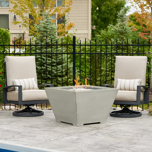 Outdoor Greatroom Co Cove Square gas fire pit bowl in use, flanked by two outdoor chairs on a patio setting.