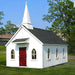 A quaint Little Cottage Chapel Playhouse with a white steeple and red doors, nestled in a green pastoral setting with a clear blue sky overhead.