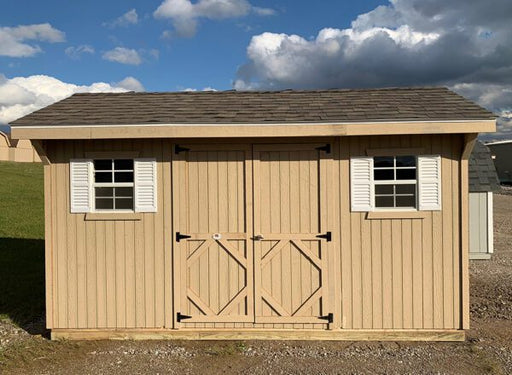 Front view of a traditional Classic Saltbox Shed with shutters, displaying the distinctive roof slope, situated on a gravel base against a cloudy sky.