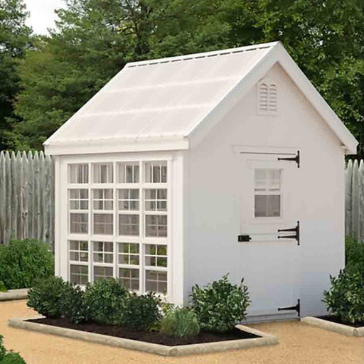 A white Colonial Greenhouse from Little Cottage Company surrounded by manicured shrubs and a gravel path.