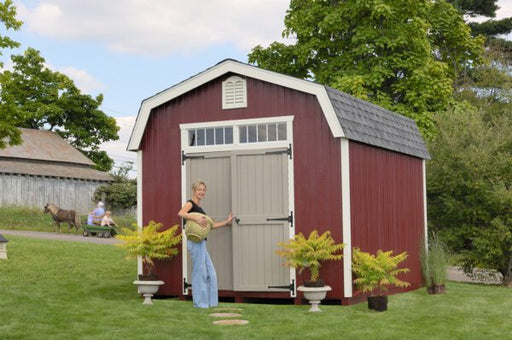 A smiling person opening the double doors of the red and beige Colonial Woodbury garden shed, with a horse-drawn cart visible in the background.