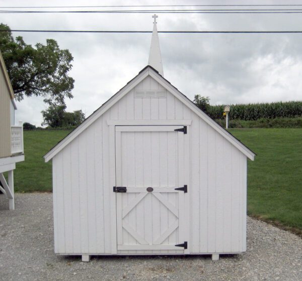 The front view of a Little Cottage Chapel Playhouse, displaying simple white architecture with a cross on the peak, set against a rural backdrop.