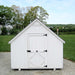 The front view of a Little Cottage Chapel Playhouse, displaying simple white architecture with a cross on the peak, set against a rural backdrop.