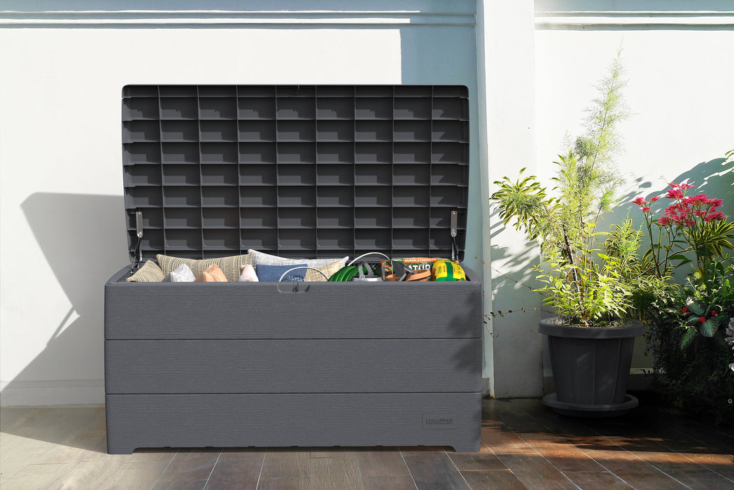 The Duramax 416 Liter/110 Gallon Outdoor Storage Box in grey, open and filled with cushions and outdoor essentials, positioned against a white wall with potted plants nearby.