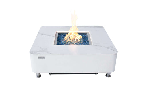 A square white marble fire pit featuring vibrant blue fire glass flames, showcasing the Elementi Plus Annecy Porcelain Top Fire Table