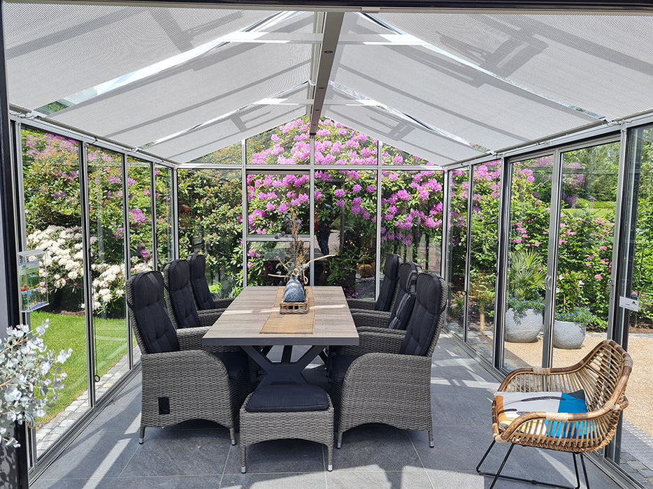 Interior of a glass greenhouse with a peaked transparent roof, furnished with a wooden dining table and comfortable chairs, surrounded by vibrant flowering plants and greenery visible through the clear panes.