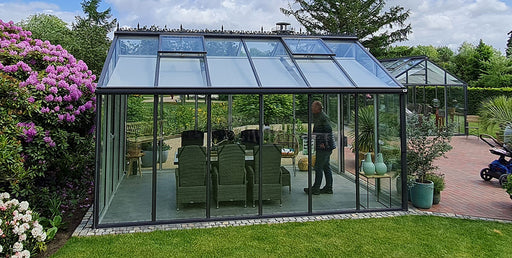 Prominent display of a glass greenhouse with a black frame, featuring a functional vent system on the roof, set against a garden with lush greenery, emphasizing the product's design and capacity for plant cultivation.
