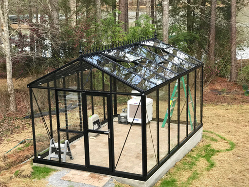 An Exaco Royal Victorian VI-34 greenhouse structure with a black aluminum frame and clear glass panels in a backyard garden.