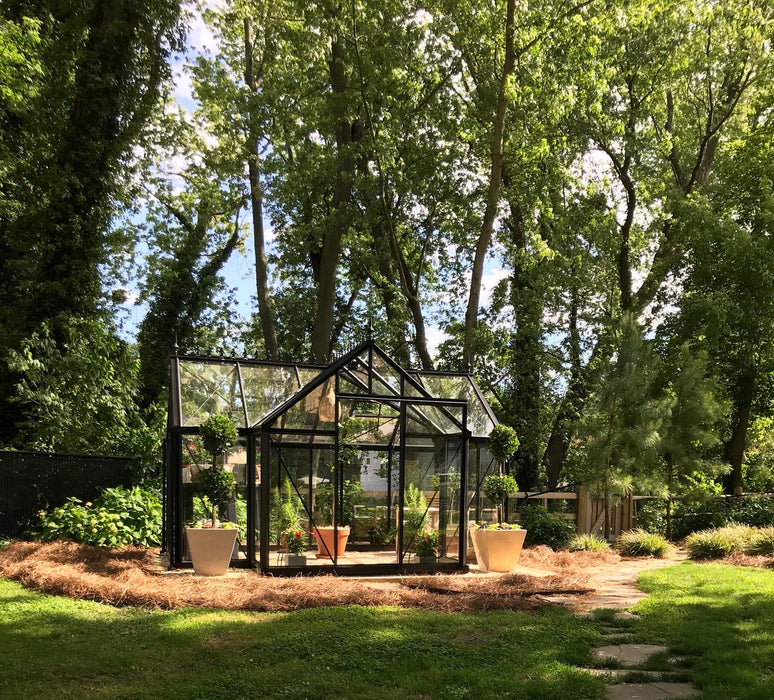 Peaceful setting of a Janssens Exaco Junior Orangerie Greenhouse, nestled in a shady garden with mature trees and a natural backdrop.