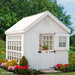 Pristine white Colonial Gable Greenhouse by Little Cottage Company nestled in a lush garden.