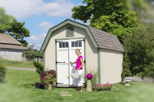 Woman standing by a beige Colonial Greenfield Shed in a vibrant garden setting from Little Cottage Company.