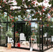 Exaco Janssens M23 modern greenhouse featuring a sloping roof, with a dog resting inside near white garden chairs.