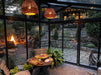 An inviting Exaco Janssens Modern Greenhouse at twilight with warm lighting, showcasing an intimate dining setup overlooking a serene garden with a fire pit.