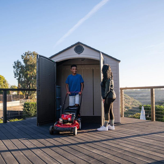 A teenager moving a red lawnmower out of the Lifetime 8 ft x 5 ft Outdoor Storage Shed while an adult observes.