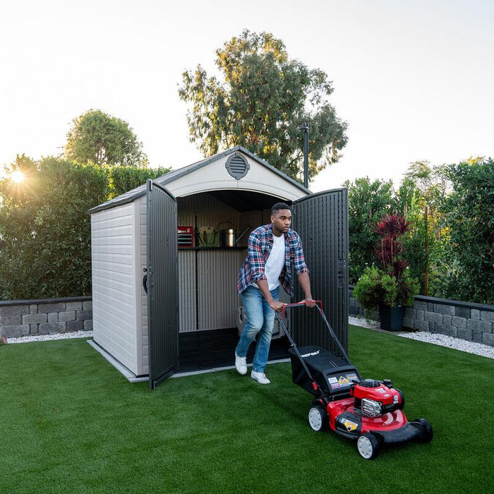 A person exiting the Lifetime 8 ft x 7.5 ft Outdoor Storage Shed with a lawn mower, demonstrating the usage and interior space of the shed.