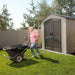 A senior woman with a wheelbarrow standing next to the Lifetime 7 x 7 ft Outdoor Storage Shed.