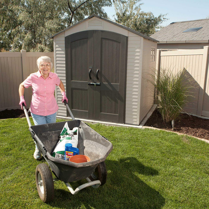  Lifetime 7x7 ft Outdoor Storage Shed in a backyard with a senior woman pushing a wheelbarrow.