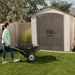 A woman walking with a wheelbarrow towards the closed Lifetime 7 x 7 ft Outdoor Storage Shed in a backyard.