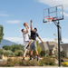 Two players in mid-air competing for a basketball near the Lifetime Adjustable In-Ground Basketball Hoop with a clear blue sky in the background.