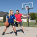 Two women playing basketball with the Lifetime Adjustable Bolt Down Basketball Hoop in the background.