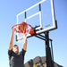 Teen boy stretching to dunk on the Lifetime Adjustable Portable Basketball Hoop with a clear blue sky in the background and a suburban house.
