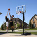 Teen boy jumping to shoot a basketball with another boy in defense, playing at a Lifetime Adjustable Basketball Hoop.
