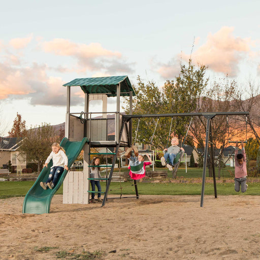 Full view of the Lifetime Adventure Clubhouse Playset, SKU 91135, in an outdoor setting at dusk with children playing on the swings and slide, with a backdrop of a residential area and a sky with clouds.