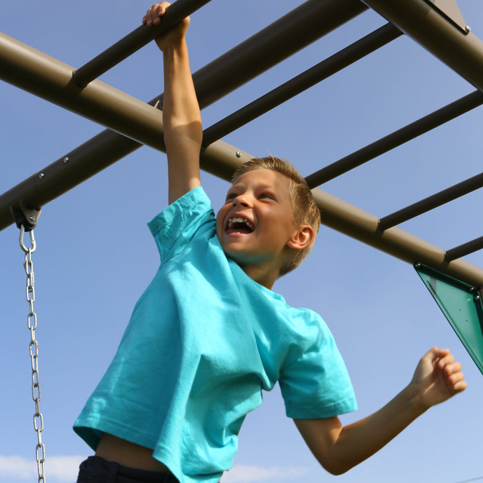 A boy in a blue shirt hanging from the monkey bars of the playset.