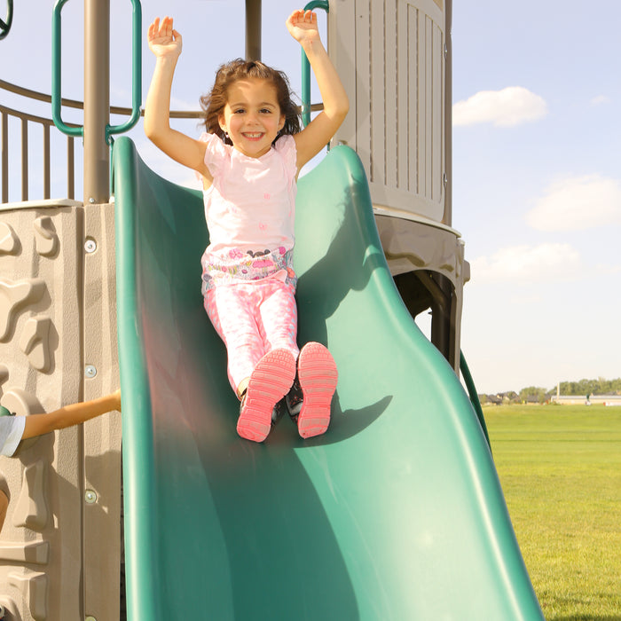 A young girl in pink sliding down the green slide of the playset.