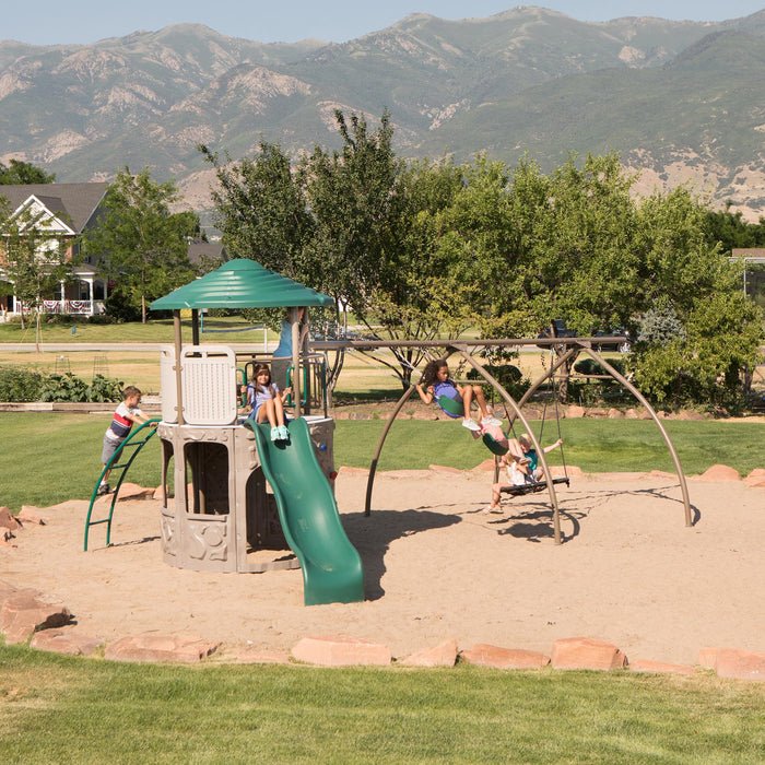 Distant view of the Lifetime Adventure Tower Playset featuring a slide and swings with children playing, set against a backdrop of trees and mountains.