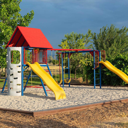 Wide-angle view of the Lifetime Big Stuff Deluxe Swing Set featuring a red clubhouse, blue and red swings, yellow slides, and a rock-climbing wall, set in a sand-covered play area.