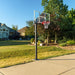 Side view of a Lifetime Crank Adjust Bolt Down Basketball Hoop installed on a grassy backyard with clear skies and a residential setting in the background.