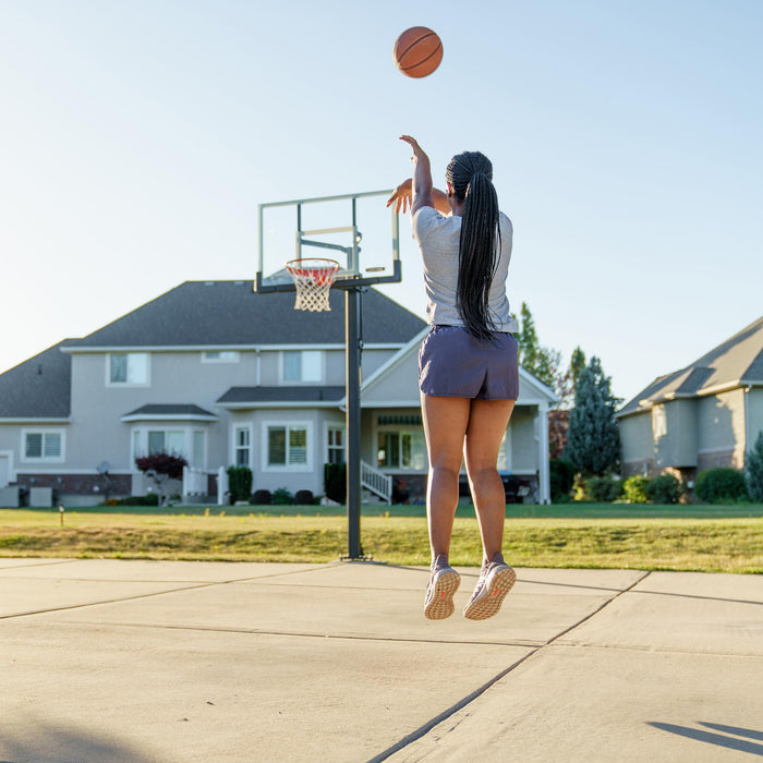 A person shooting a basketball towards a Lifetime Crank Adjust Bolt Down Basketball Hoop with a clear backboard in a residential driveway.