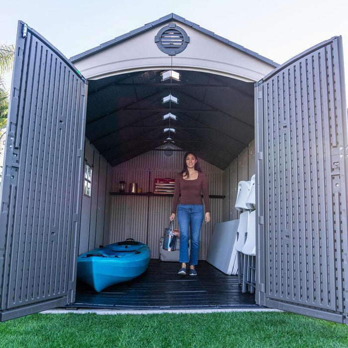 Woman walking out of the Lifetime 8 Ft. x 15 Ft. Outdoor Storage Shed with an open door revealing interior and kayak.