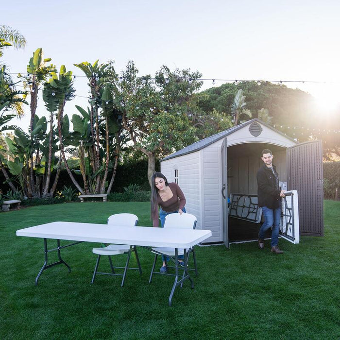 Two people setting up a folding table and chairs outside of the Lifetime 8 Ft. x 15 Ft. Outdoor Storage Shed in a garden.
