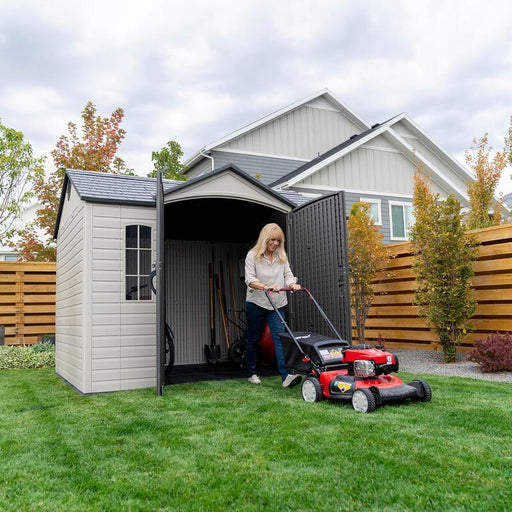 Woman pushing a lawnmower out of the Lifetime 10 x 8 ft. Outdoor Storage Shed in a backyard setting.