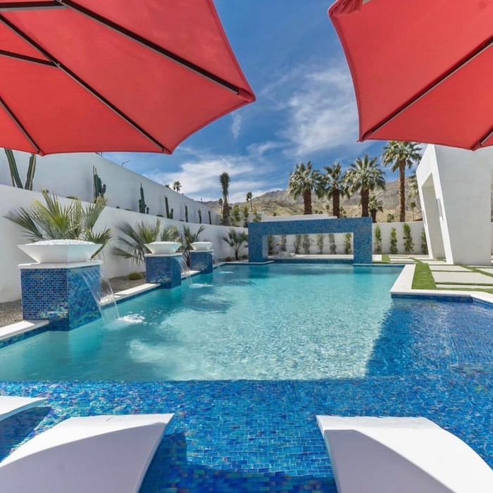 Luxurious poolside view with a square GFRC concrete fire bowl as a central feature, surrounded by vibrant red umbrellas and a serene blue pool.