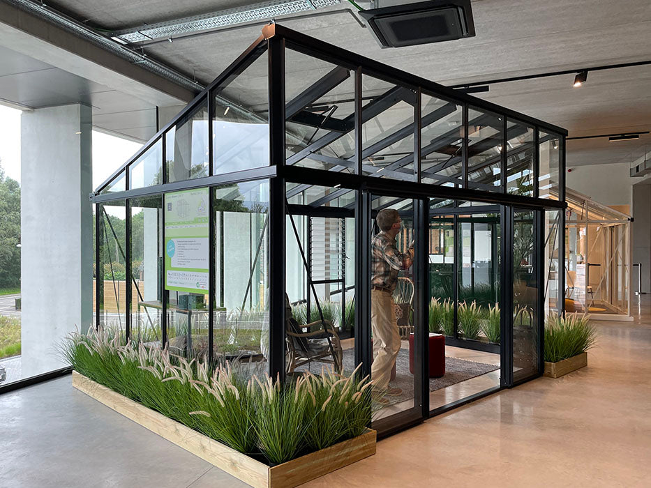 Inside view of a modern Exaco Janssens M23 greenhouse, showing a person tending to plants, surrounded by elegant black framing.